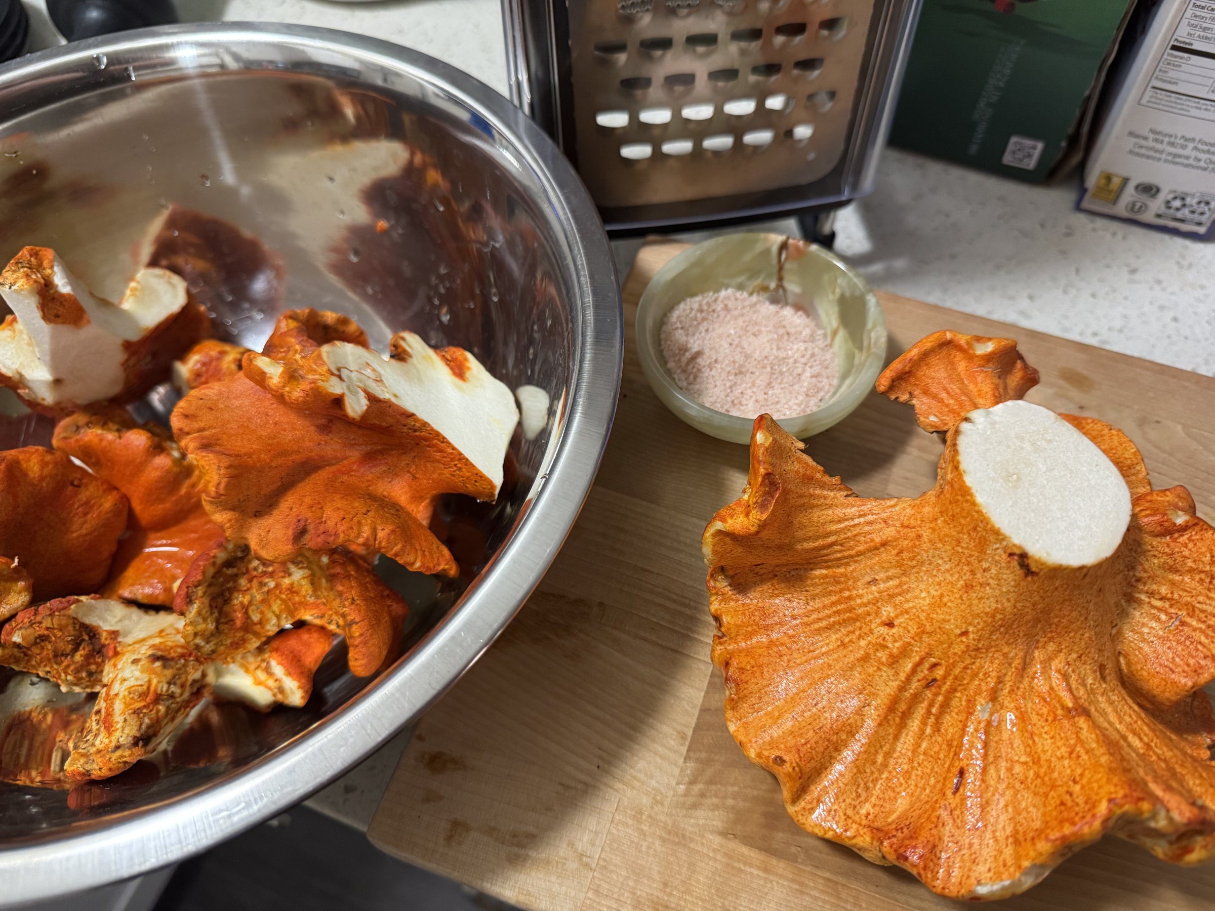 A large lobster mushroom cut open on a wooden board, bright orange crust outside and solid white flesh inside, with a bowl of cleaned pieces beside it